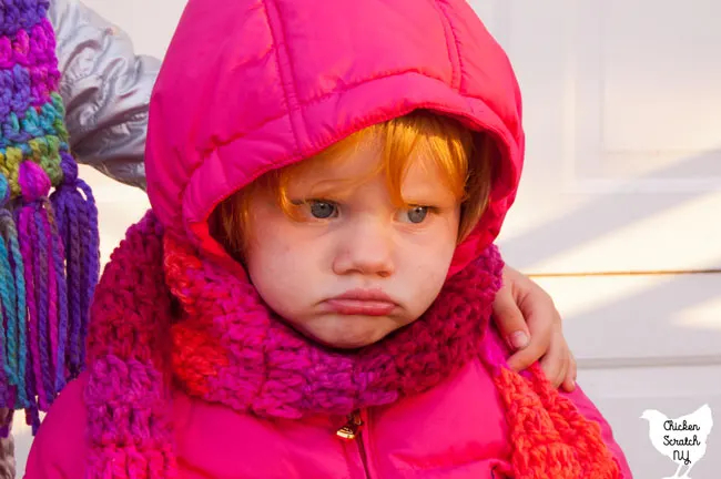 grumpy little blonde girl in puffy pink winter coat with hand crocheted pink and orange scarf