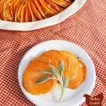 red deep dish pie plate filled with sliced sweet potatoes baked in a maple sage cream sauce on a red checkered cloth with a small white plate with a fan of sweet potato slices and a fresh sage leaf