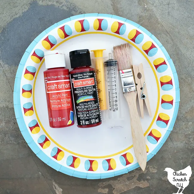 paper plate holding two colors of red paint, a medcine syringy, a paint brush and a clothes pin used to make a bloody Halloween apron