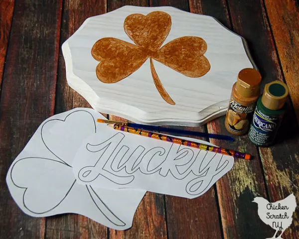 white wooden sign handpainted with a metallic gold shamrock and two paper stencils and green and gold paint