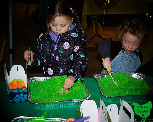 little boy with curly hair and little girl in winter coat using paintbrush to dig through green play sand for dinosaur bones