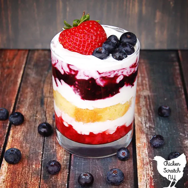 glass jar on a wooden table layered with strawberries, whipped cream, lemon pound cake and blueberries topped with fresh blueberries and a strawberry