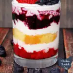 glass jar on a wooden table layered with strawberries, whipped cream, lemon pound cake and blueberries topped with fresh blueberries and a strawberry