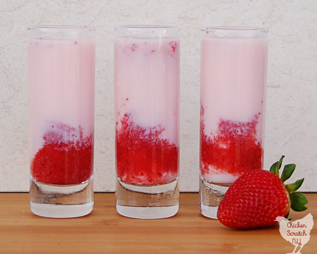 three shooter glasses filled with pink opaque liquid an red strawberry puree on a wooden cutting board with a red, ripe strawberry