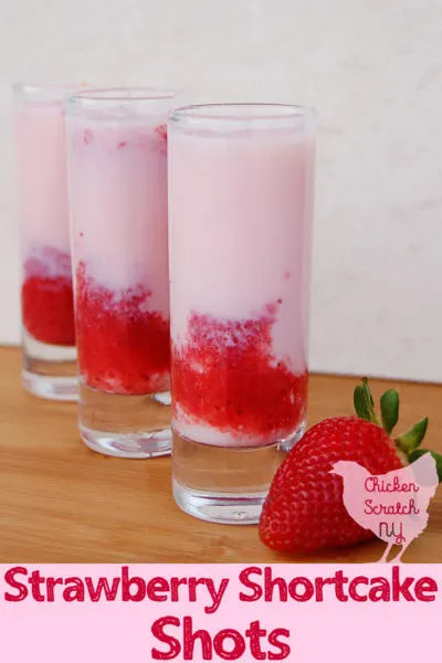 three shooter glasses filled with pink opaque liquid an red strawberry puree on a wooden cutting board with a red, ripe strawberry