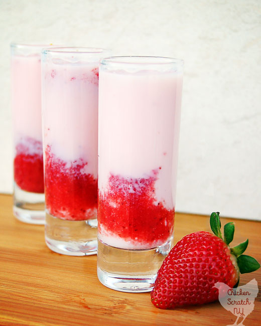 three shooter glasses filled with pink opaque liquid an red strawberry puree on a wooden cutting board with a red, ripe strawberry