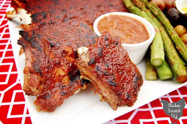rhubarb BBQ sauce covered ribs on a white plate on a red and white table cloth with a white dish filled with more BBQ sauce next to broiled asparagus