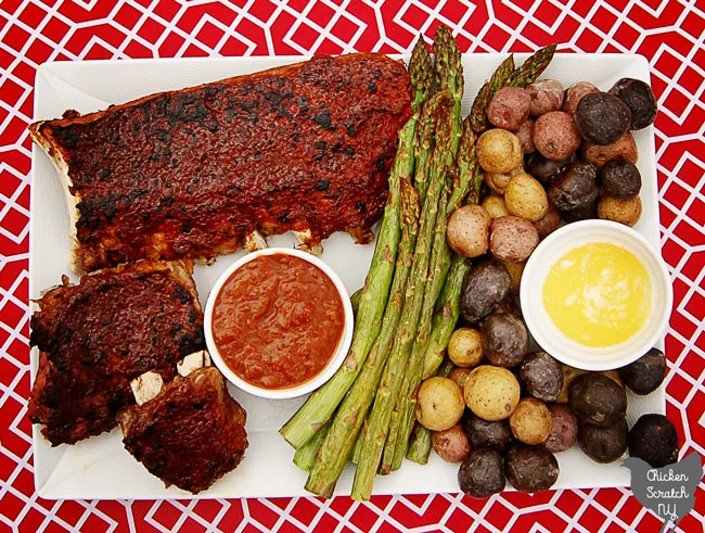 rhubarb BBQ sauce covered ribs on a white late on a red and white table cloth with a white dish filled with more BBQ sauce, broiled asparagus and red white and blue salt potatoes with a white dish of melted butter