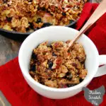 baked oatmeal with rhubarb, pecans and blueberries in a cast iron skillet with a scoop in a white cup with a copper spoon, on a red towel and rhubarb stalks on a grey stained wooden board