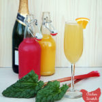 champagne, orange and rhubarb juice in flip top bottles, champagne glass filled with rhubarb mimosa with orange garnish with two stalks of rhubarb