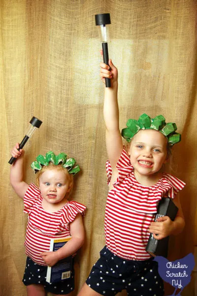 two little girls in red, white and blue outfits each wearing a handmade green statue of liberty crown holding a book and a solar lantern