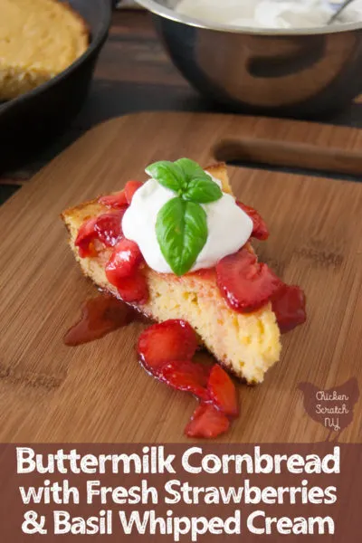 buttermilk cornbread topped with fresh strawberries and whipped cream with bail leaf on wooden cutting board with cast iron pan of cornbread and metal bowl of whipped cream in the back