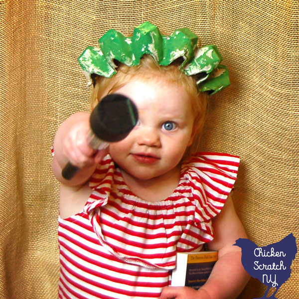 little girl in red, white and blue outfit wearing a handmade green statue of liberty crown holding a book and a solar lantern