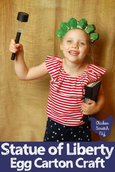 little girl in red, white and blue outfit wearing a handmade green statue of liberty crown holding a book and a solar lantern