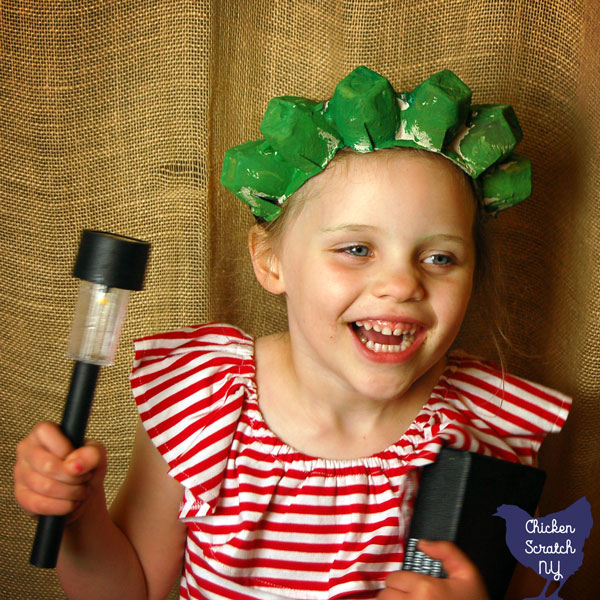 little girl in red, white and blue outfit wearing a handmade green statue of liberty crown holding a book and a solar lantern