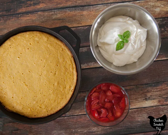 buttermilk cornbread topped with fresh strawberries and whipped cream with bail leaf on wooden cutting board with cast iron pan of cornbread and metal bowl of whipped cream in the back