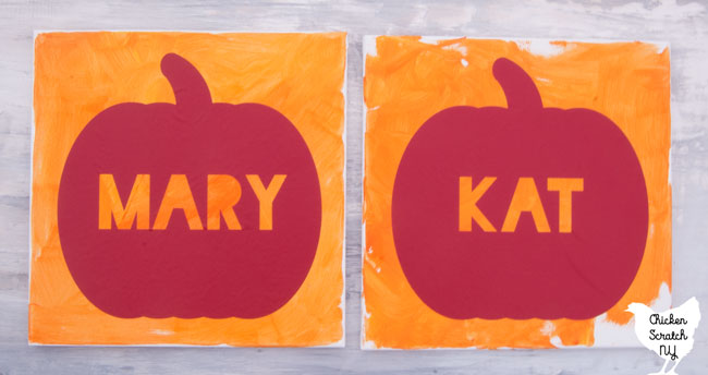 two 12x12 canvases painted with varying shades of orange craft paint with a red vinyl stencil on each one with the name MARY and one with the name KAT