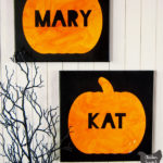 white wall with two hanging black canvases with orange pumpkins and names in black block letters, one says MARY and the other says KAT with creep black branches in the corner