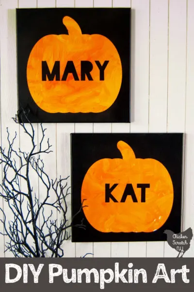 white wall with two hanging black canvases with orange pumpkins and names in black block letters, one says MARY and the other says KAT with creep black branches in the corner