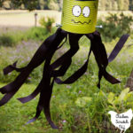 tin can turned into DIY Frankenstein Windsock with paint, screws and ribbon hanging in a tree with the black ribbon streamers blowing in the breeze