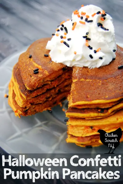 stack of Halloween confetti pumpkin pancakes filled with freamy pumpkins and black and orange sprinkles on a spiderweb frosted glass plate topped with whipped cream and more sprinkles