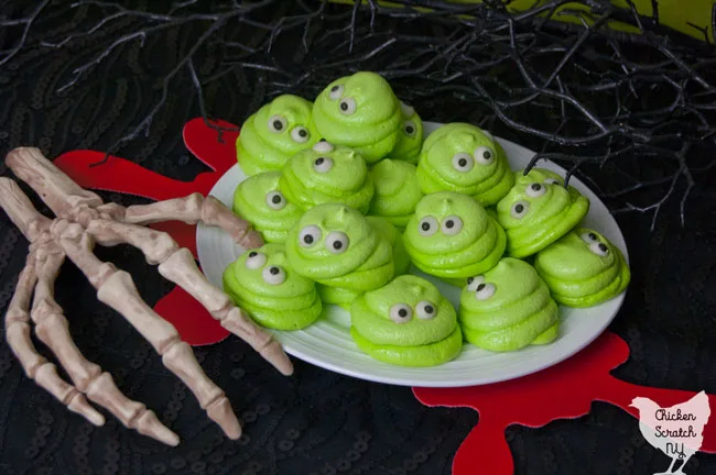 white plate full of lime green meringue cookies with eyes that look like Blobby from Hotel Transylvania on a black surface with red blood splatter coasters and a skeleton hand