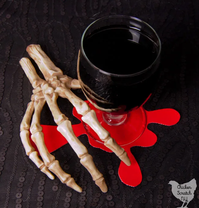 skeleton hand holding glass of dark liquid on top of bright red blood spatter coaster