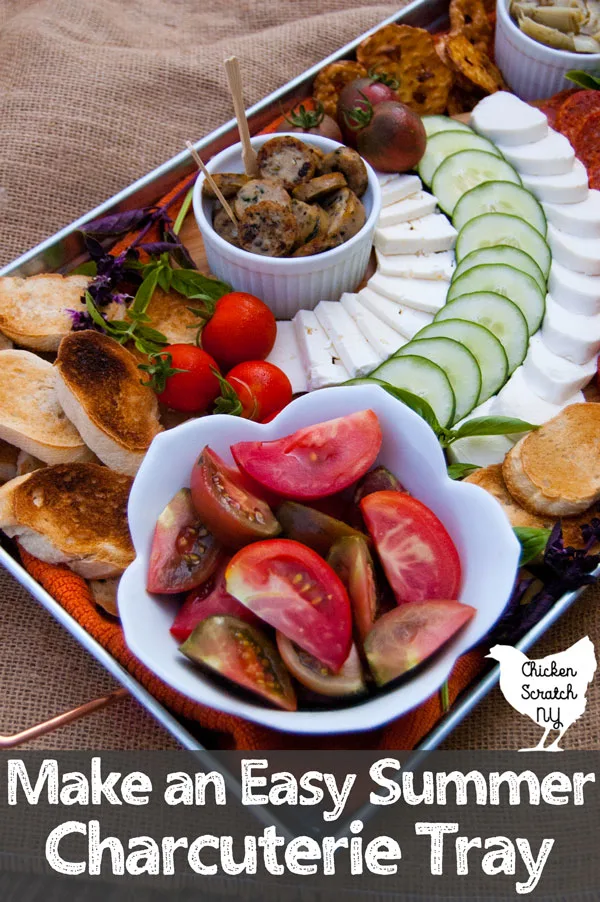 bowl of sliced red and purple tomatoes on a metal tray with a cutting board with mozzarella cheese, feta cheese, pepperoni, cherry tomatoes and toasted bread with fresh basil