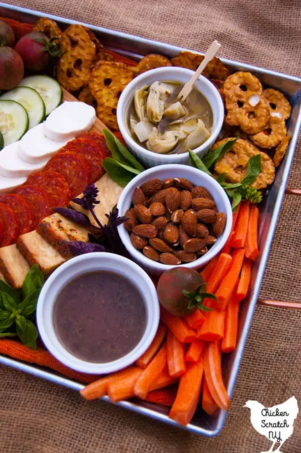 a metal tray with a cutting board with mozzarella cheese, feta cheese, pepperoni, cherry tomatoes, carrot sticks and toasted bread, pretzel thins, cucumber slices, marinated artichoke hearts, almonds and vinaigrette with fresh basil
