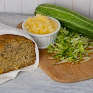 homemade sliced pineapple zucchini bread wrapped in a white tea towel in front of a cutting board with shredded zucchini and crushed pineapple in a white dish