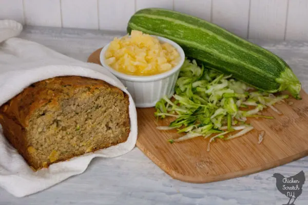 homemade sliced pineapple zucchini bread wrapped in a white tea towel in front of a cutting board with shredded zucchini and crushed pineapple in a white dish