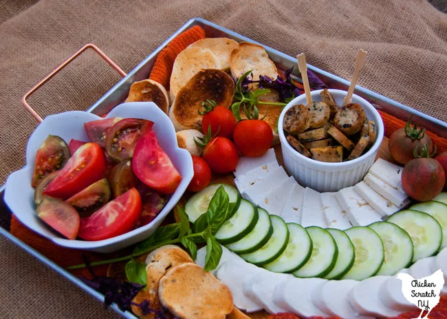 bowl of sliced red and purple tomatoes on a metal tray with a cutting board with mozzarella cheese, feta cheese, pepperoni, cherry tomatoes and toasted bread with fresh basil