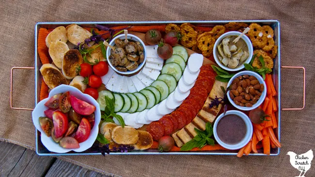 bowl of sliced red and purple tomatoes on a metal tray with a cutting board with mozzarella cheese, feta cheese, pepperoni, cherry tomatoes and toasted bread, pretzel thins, cucumber slices, marinated artichoke hearts, almonds and vinaigrette with fresh basil