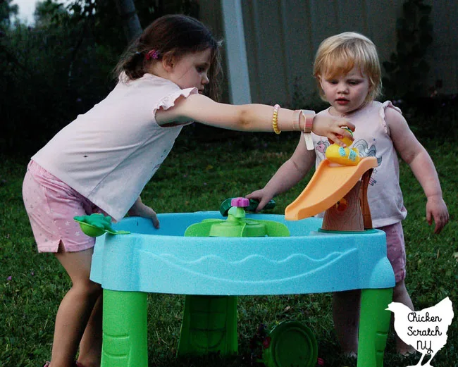 two little girls in pink shorts and pink shirts playing with a blue water table with rubber ducks and a frog