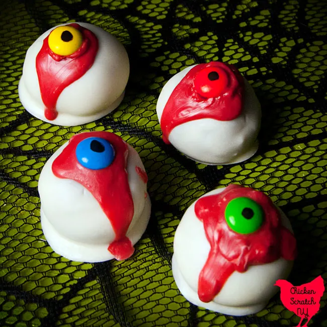 four red velvet oreo truffles dipped in white candy melts with a red drizzle and a blue, yellow, green and orange m&m eye with a black pupil on a green surface with a black spiderweb pattern