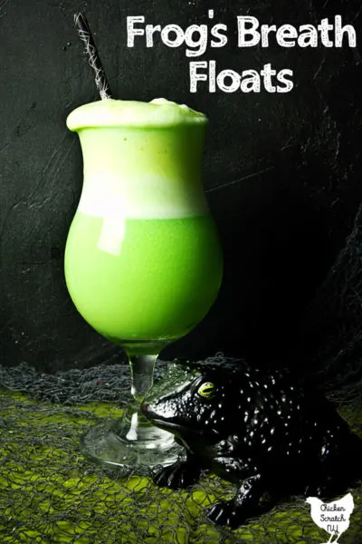 lime sherbet, pineapple and lemon lime soda float in a tall ice cream glass with a black frog in the bottom right corner on a green surface with grey creepy netting and a black background
