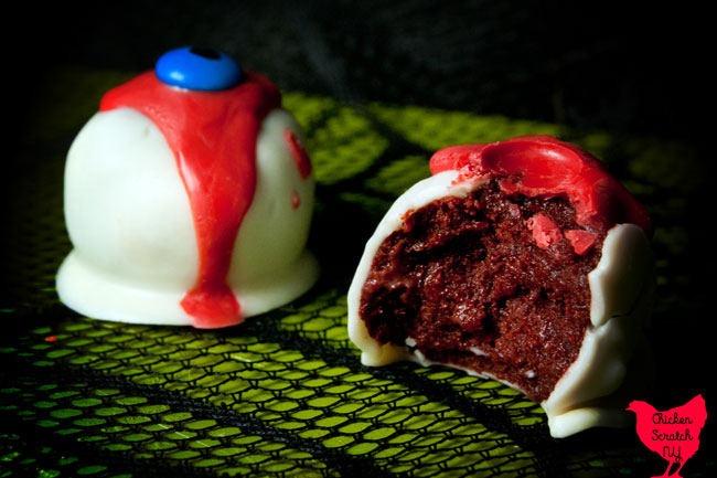 two red velvet oreo truffles dipped in white candy melts with a red drizzle and a blue m&m eye with a black pupil with one bitten open to show the red inside on a green surface with a black spiderweb pattern