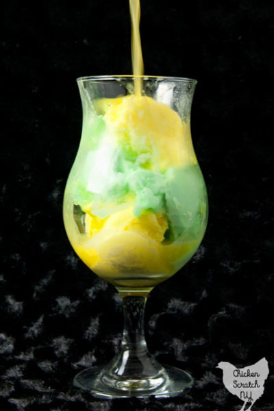 pineapple juice pouring over clear ice cream glass filled with lemon and lime sherbet against a black background