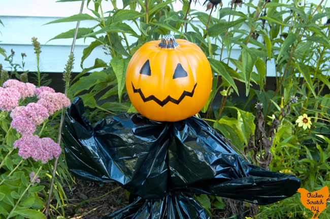 garden stake turned onto a Halloween garden sculpture with a small fake pumpkin, trash bag and zip ties standing in a late summer garden