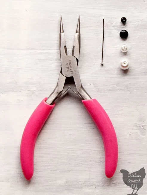 pair of jewelry pliers, a large white pearl, a medium white pearl, a flattened black bead and a round black bead with a silver head pin