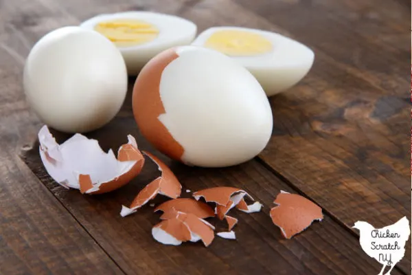 Three hard boiled eggs, one fully shelled and cut in half, two partial peeled with brown egg shell pieces on a wooden surface