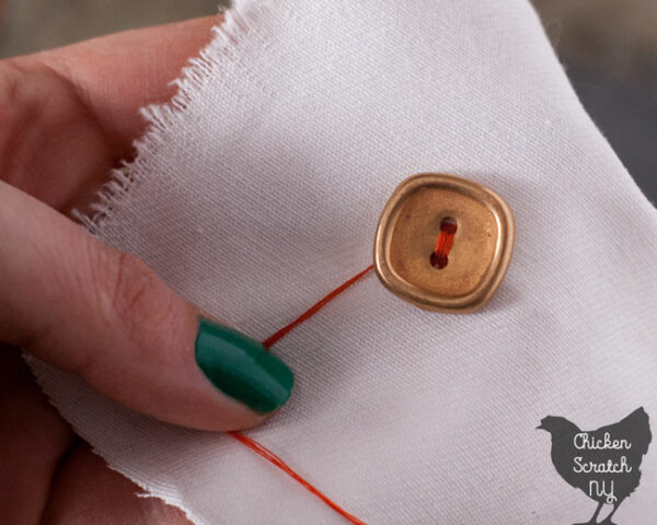 hand sewing a flat button to while fabric with orange thread wrapping around the stitches to create a shank