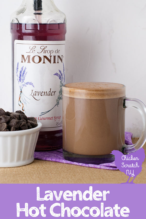 clear glass mug filled with lavender hot chocolate on a rough burlap surface with a bottle of lavender syrup and a ramekin of chocolate chips