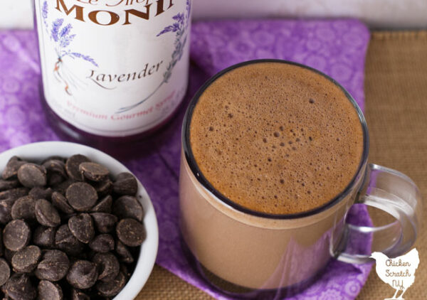 clear glass mug filled with lavender hot chocolate on a rough burlap surface with a bottle of lavender syrup and a ramekin of chocolate chips