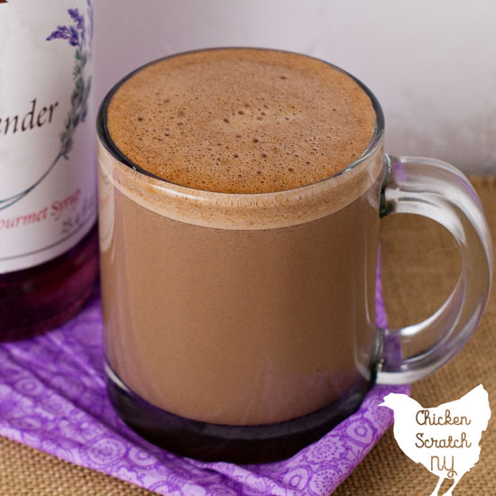 clear glass mug filled with lavender hot chocolate on a rough burlap surface with a bottle of lavender syrup