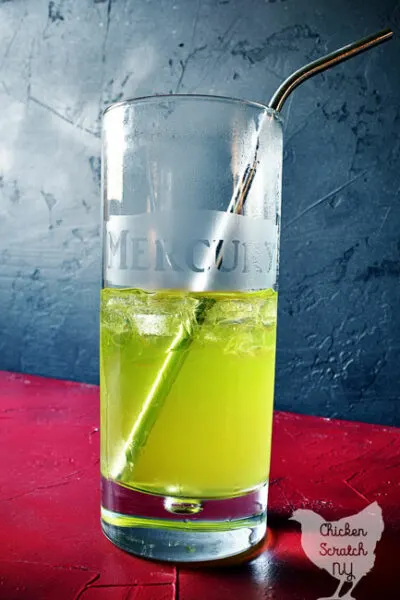 tall glass with mercury etched on the front filled with green liquid and a metal straw