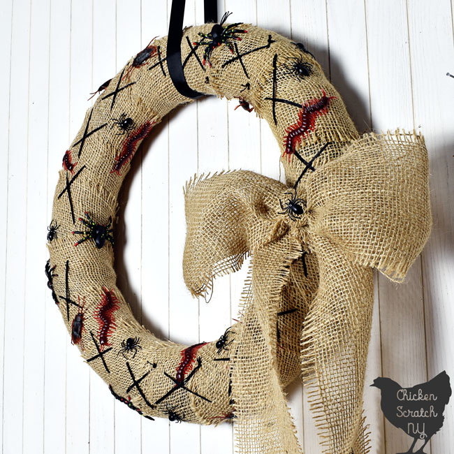 close up of burlap covered Nightmare Before Christmas Wreath with fake bugs and spiders with a burlap bow hanging on a white wall with a black ribbon