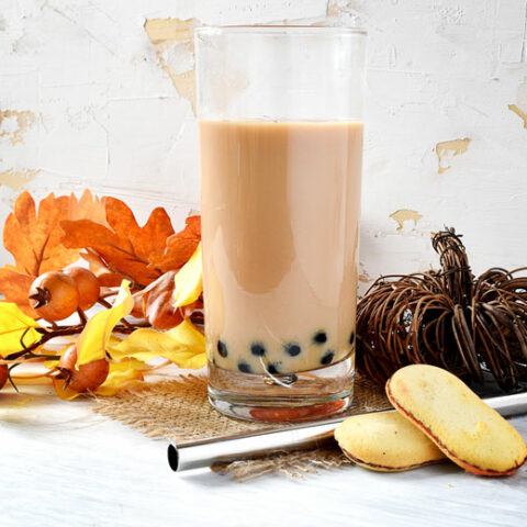 tall glass filled with cramy tan tea with black boba pearls, two milano cookies, a wicker pumpkin, fake leaves and a silver bubble tea straw