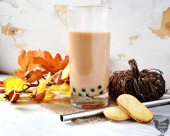 tall glass filled with cramy tan tea with black boba pearls, two milano cookies, a wicker pumpkin, fake leaves and a silver bubble tea straw