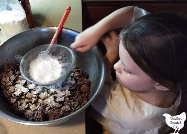 little girl with powdered sugar in a sifter with a large metal bowl filled with muddy buddies